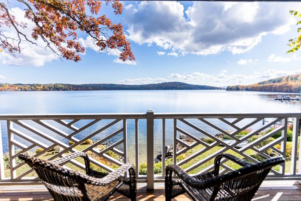 Two wicker chairs on a deck facing a serene lake under a partly cloudy sky with autumn trees in the foreground.