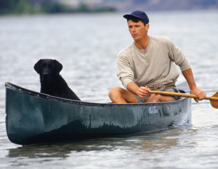 A man is paddling a canoe on a body of water with a black dog sitting at the front of the canoe.