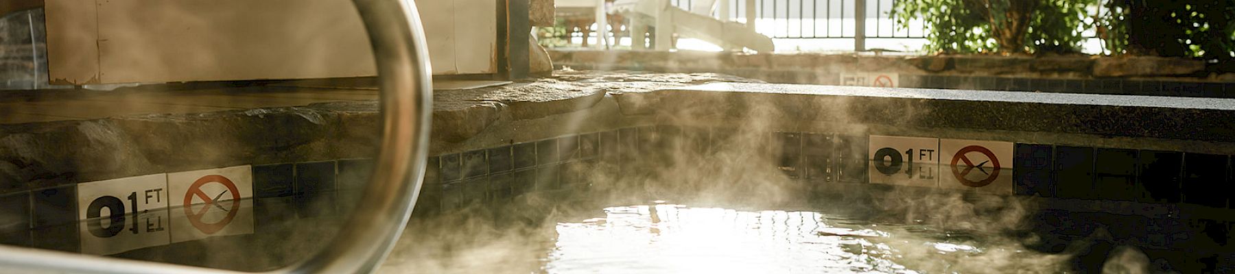 There is a steamy hot tub next to a building with chairs and plants seen beyond a railing on a sunny day.