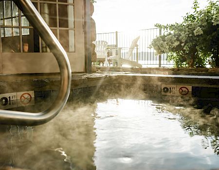 There is a steamy hot tub next to a building with chairs and plants seen beyond a railing on a sunny day.