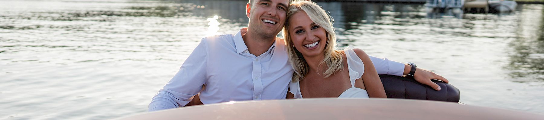 A couple smiles while sitting on a boat, with a picturesque waterfront scene and buildings in the background during daylight.