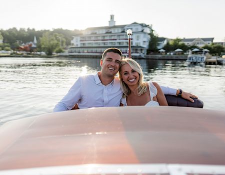 A couple smiles while sitting on a boat, with a picturesque waterfront scene and buildings in the background during daylight.