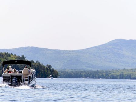 A pontoon boat with people onboard moves across a lake with mountainous scenery in the background. The sky is clear and the water is calm.