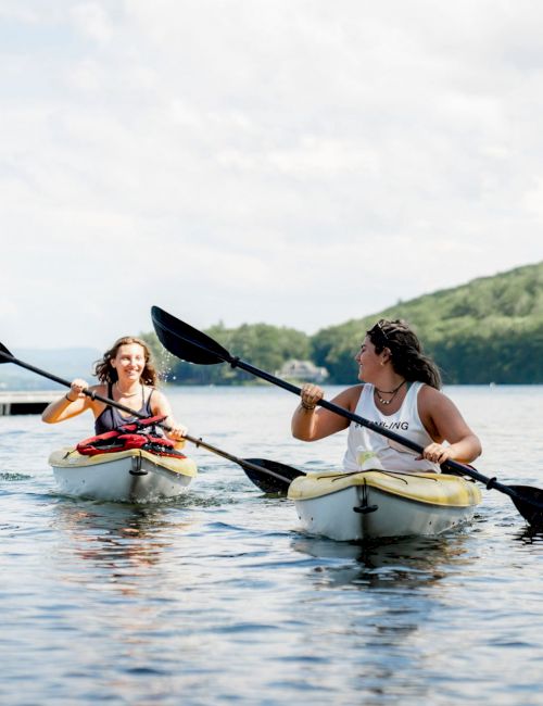 Two people are kayaking on a calm lake surrounded by green hills and a floating dock in the background.