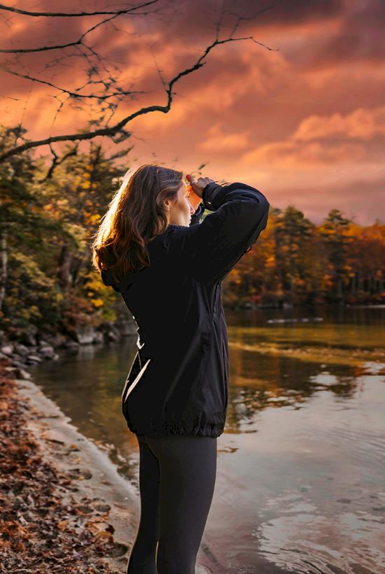 A person stands by a lake, wearing a black jacket, taking photos of a vibrant, colorful sunset with trees reflecting on the water.