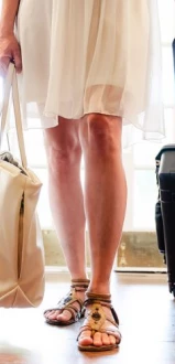 A woman dressed in a white dress carries a beige bag and a suitcase with wheels, standing in a sunlit room.