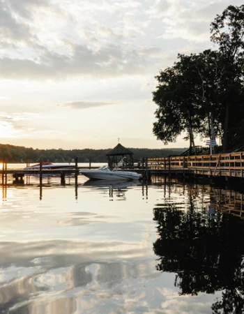 A serene lakeside view at sunset with a wooden pier, boats, and trees reflecting on the calm water, creating a peaceful and picturesque scene.