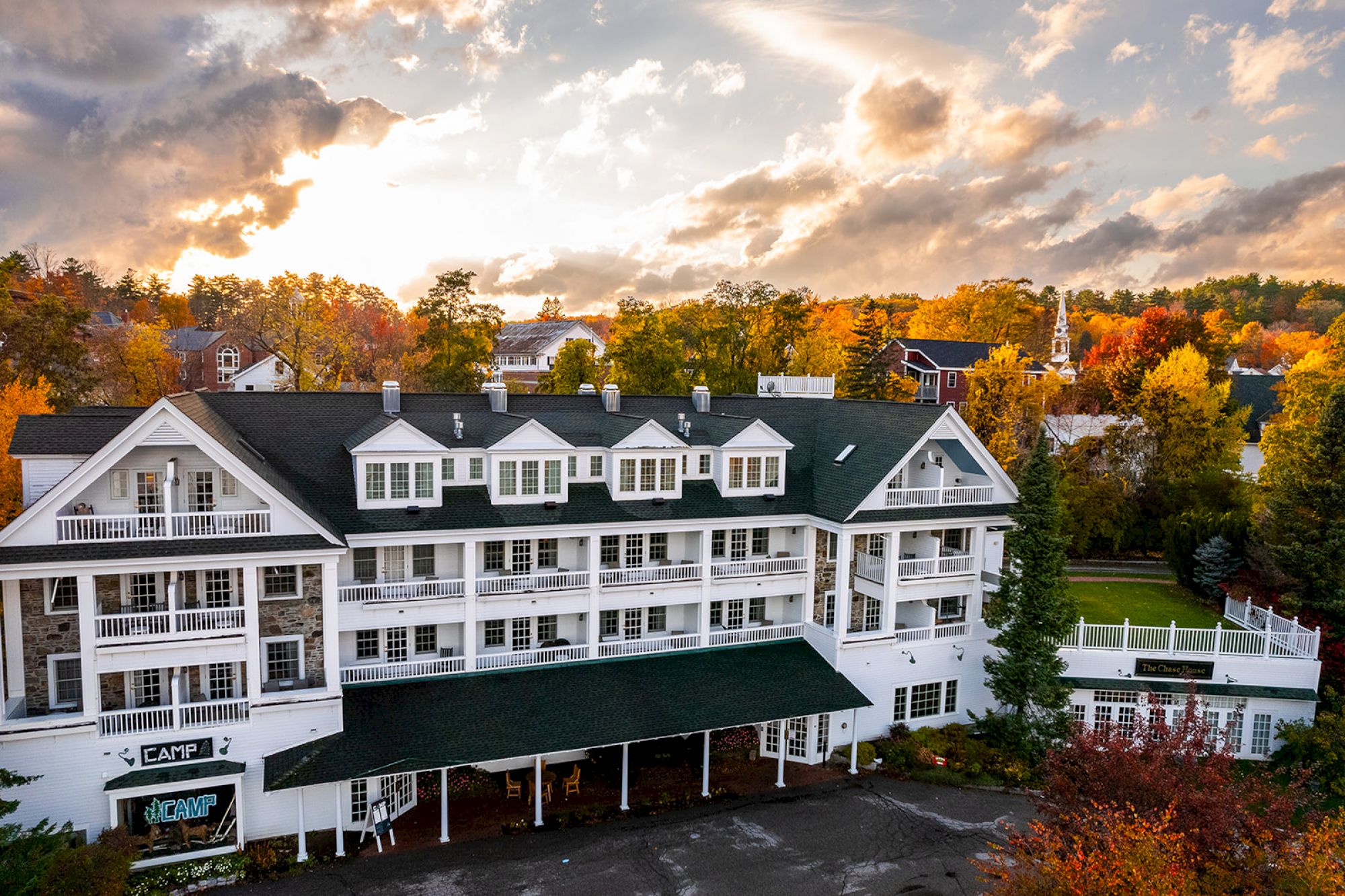 A large white building with a green roof sits amidst colorful autumn foliage under a partly cloudy sky at sunset.