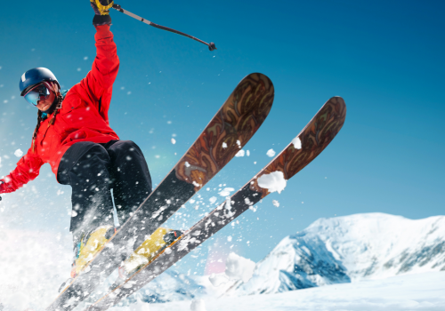 A skier in a red jacket and black pants is jumping in the air on snowy slopes with mountains in the background and a clear blue sky.