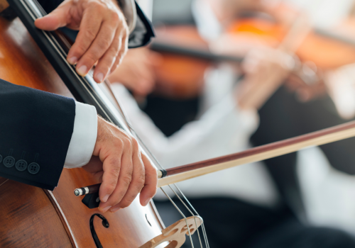 The image shows musicians playing string instruments: a close-up of hands playing a cello and blurred background of violinists.