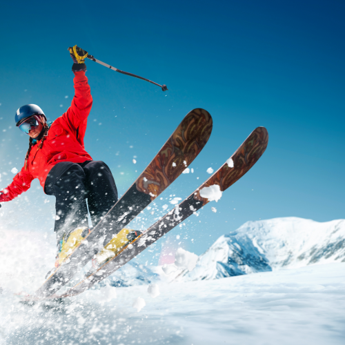 A skier in a red jacket is airborne, performing a jump with snow-capped mountains in the background, under a clear blue sky.