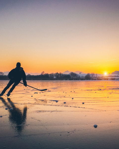 A person is ice skating on a frozen lake at sunset, with mountains in the background and the reflection of the scene visible on the ice.