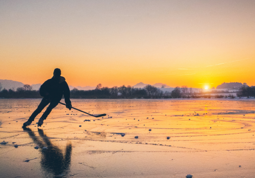 A person is ice skating on a frozen lake at sunset, with mountains in the background and the reflection of the scene visible on the ice.