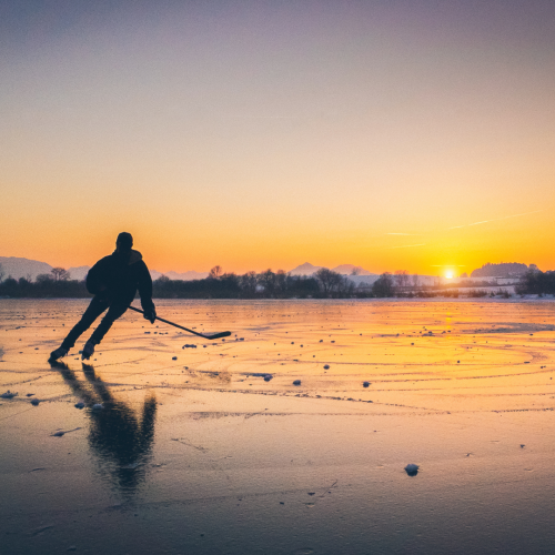 A person is ice skating on a frozen lake at sunset, with mountains in the background and the reflection of the scene visible on the ice.