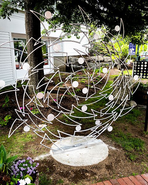 The image shows a metal sculpture with abstract designs, situated outdoors near a brick pathway and surrounded by small plants and flowers.