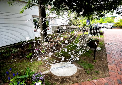 The image shows a metal sculpture with abstract designs, situated outdoors near a brick pathway and surrounded by small plants and flowers.