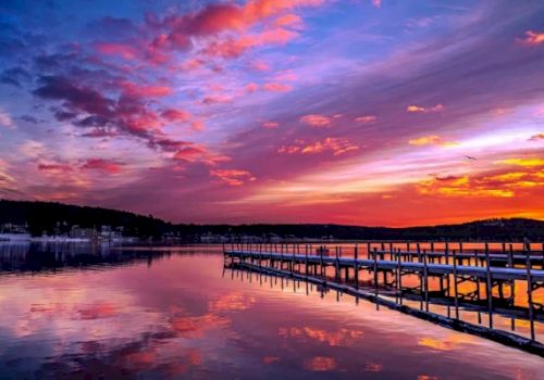 A serene lake with a wooden dock extending into the water, under a vibrant sunset sky with hues of pink, purple, and orange, ending the sentence.