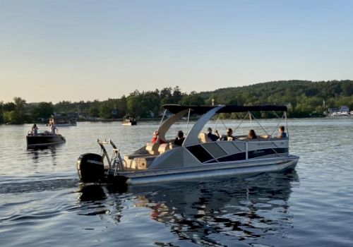 A pontoon boat filled with people is cruising on a calm lake with a distant shoreline and other boats in the background during sunset.