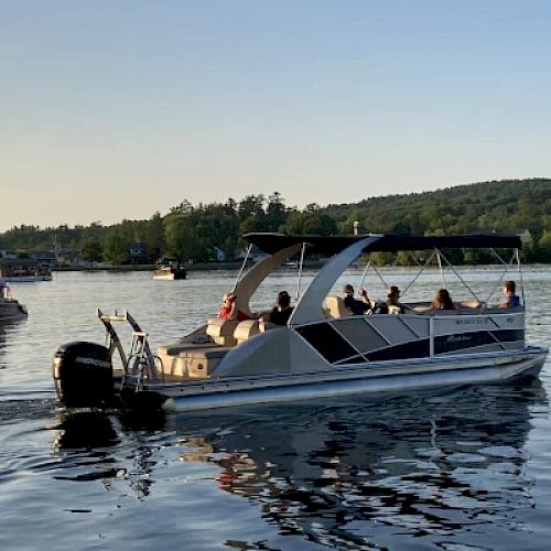 A pontoon boat filled with people is cruising on a calm lake with a distant shoreline and other boats in the background during sunset.