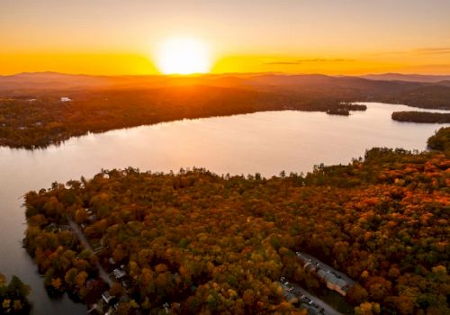 A stunning sunset over a large lake, surrounded by dense forests with autumn foliage and hills in the background, creating a serene landscape.