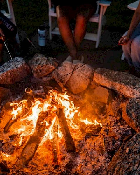 A group of people sitting around a campfire, roasting marshmallows with sticks, surrounded by rocks and seated in chairs.