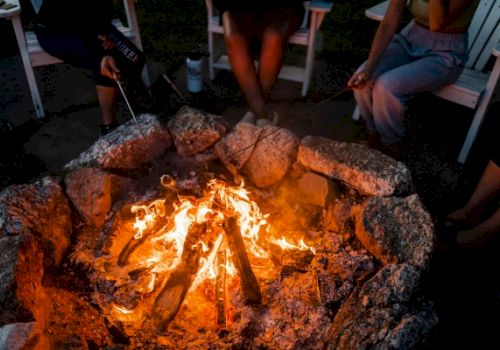 A group of people sitting around a campfire, roasting marshmallows with sticks, surrounded by rocks and seated in chairs.