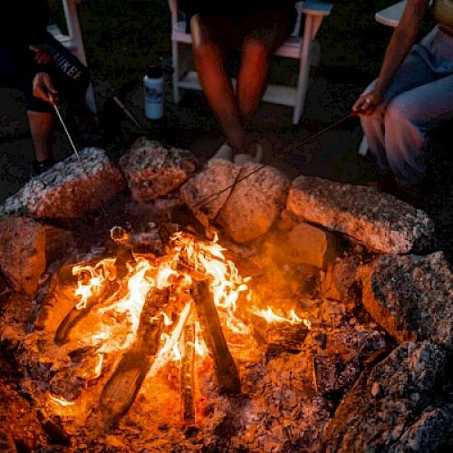 A group of people sitting around a campfire, roasting marshmallows with sticks, surrounded by rocks and seated in chairs.