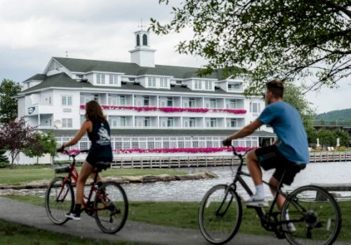 Two people are riding bikes on a path near a waterfront with a large white building adorned with pink flowers in the background.