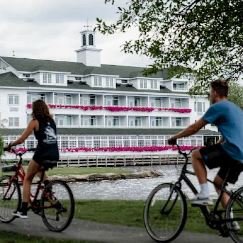 Two people are riding bikes on a path near a waterfront with a large white building adorned with pink flowers in the background.