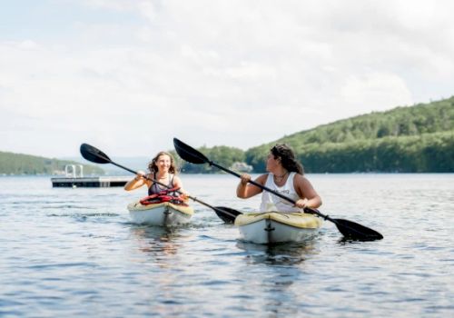 Two people kayaking on a lake with lush green hills in the background, both paddling with black oars and enjoying the water.