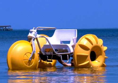 A yellow water tricycle with large buoyant wheels is floating on a calm body of water under a clear blue sky.