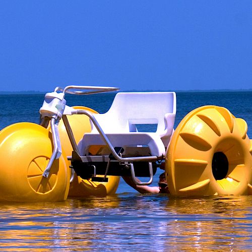 A yellow water tricycle with large buoyant wheels is floating on a calm body of water under a clear blue sky.