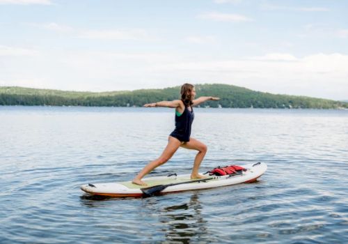 A person is doing a yoga pose on a paddleboard in the middle of a calm lake, with green hills and a blue sky in the background.