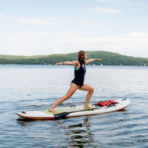 A person is doing a yoga pose on a paddleboard in the middle of a calm lake, with green hills and a blue sky in the background.