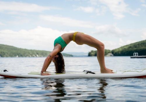 A person performing a backbend pose on a paddleboard in a calm water setting with hills and trees in the background.