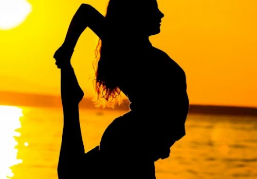 A silhouetted person is performing a yoga pose by a body of water during sunset, with their leg held up behind them in a graceful stretch.