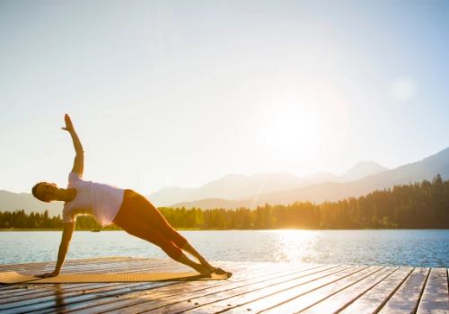 A person performs a side plank yoga pose on a wooden deck by a lake with mountains and trees in the background, under a clear sky at sunrise.