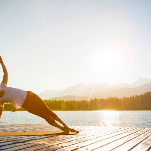 A person performs a side plank yoga pose on a wooden deck by a lake with mountains and trees in the background, under a clear sky at sunrise.