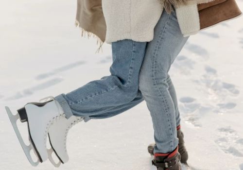 Two people are standing on ice; one person is wearing ice skates while the other person is wearing boots, both are dressed warmly.