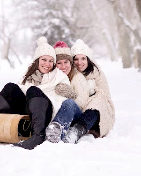 Three people in winter clothing are sitting together on the snow with a wooden sled nearby, smiling and enjoying the snowy environment.