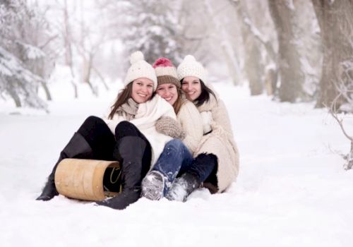 Three people in winter clothing are sitting together on the snow with a wooden sled nearby, smiling and enjoying the snowy environment.