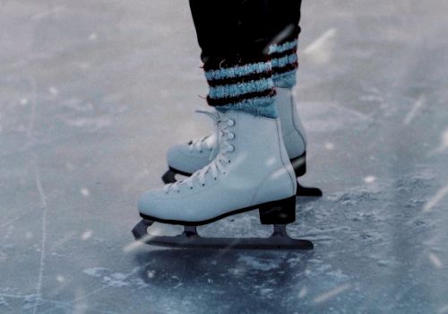 The image shows a close-up of someone's feet wearing white ice skates on an ice rink, with snow falling lightly around them.