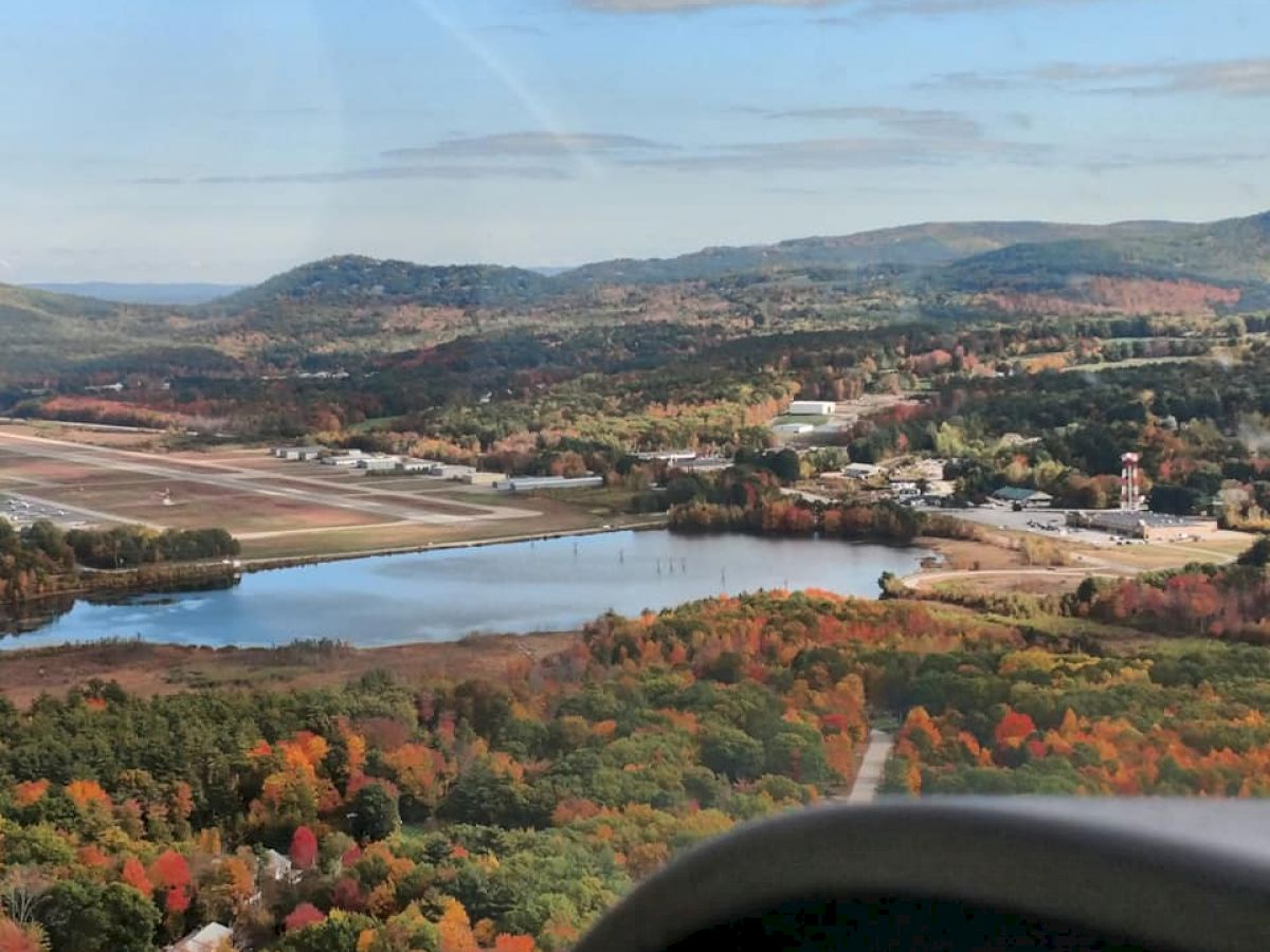 A scenic aerial view showcasing a small town surrounded by autumn-colored trees, a lake reflecting the sky, and distant rolling hills under a partly cloudy sky.