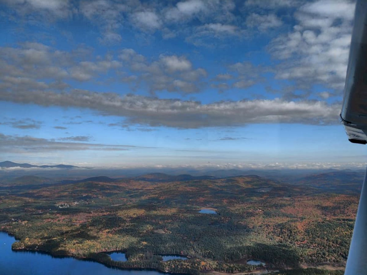 Aerial view of a scenic landscape with forests, small lakes, and distant mountains under a partly cloudy sky, wing of an airplane visible.