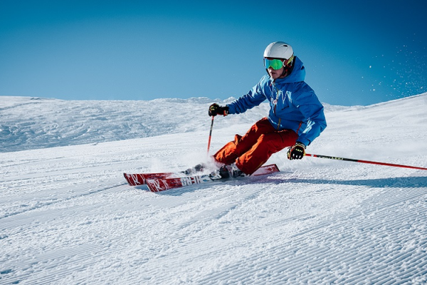 A person skiing down a snowy slope, wearing a blue jacket, red pants, and a white helmet with goggles, under a clear blue sky.