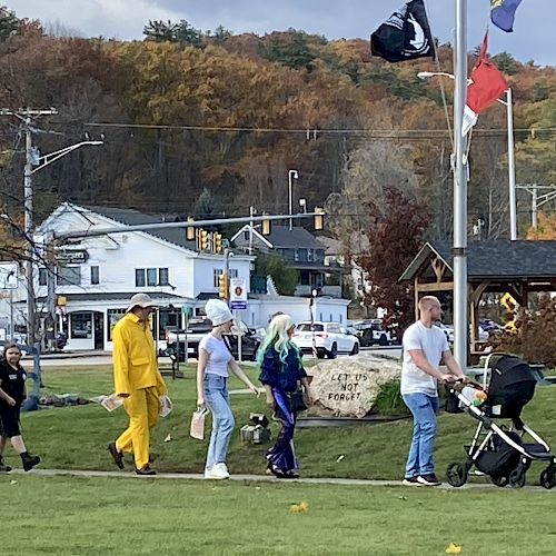 A group of people in costumes walking down a grassy path, with a small town and autumn trees in the background.