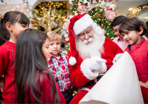 A group of children gather around Santa Claus, who is seated and holding a long scroll, in a festive Christmas setting with a decorated tree.