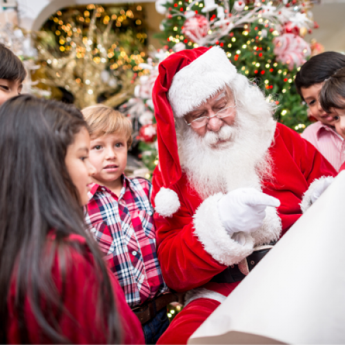 A group of children gather around Santa Claus, who is seated and holding a long scroll, in a festive Christmas setting with a decorated tree.