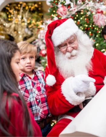 A person in a Santa costume is surrounded by children, holding a list. A decorated Christmas tree is in the background.