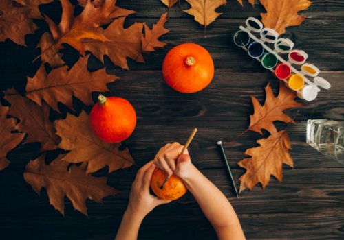 A table with pumpkins, autumn leaves, and paints; a person is painting a pumpkin with a brush, evoking a fall crafting vibe.